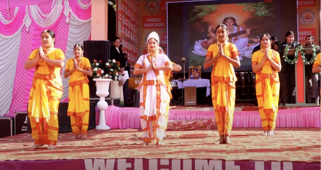Students in yellow costumes perform in front of a Saraswati backdrop
